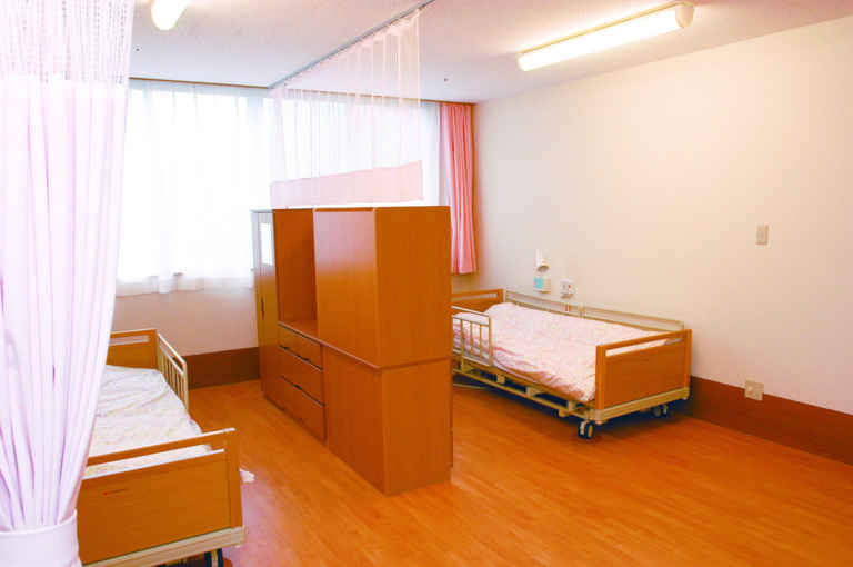 木製仕切り壁で分けられた介護施設の二人部屋。医療用ベッドと採光窓が配置。